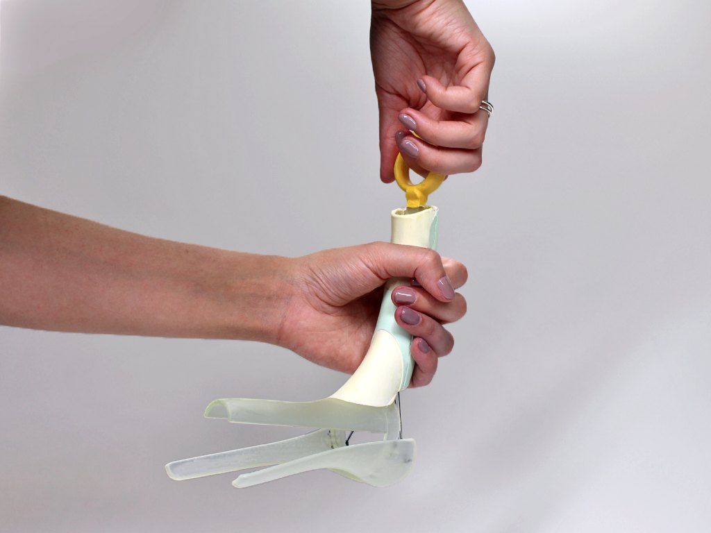 A functional model featuring the speculum in the open position. The handle comfortably fits the patient’s fingers and is pulled upwards to open. The pull handle of the device allows the handle to run along a rail and lock into the appropriate opening diameter (this is unique from person to person). This pull handle is attached to silicone elastic bands to open the blades of the speculum. The blades are attached to the main body via four hinges.